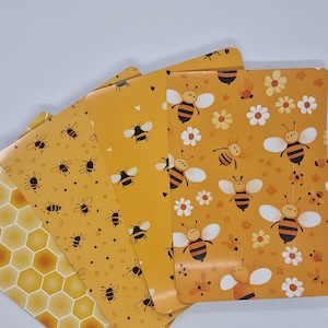 Op de afbeelding: Een stapel gele notitieboekjes met bijen- en honingraatpatronen. De notitieboekjes hebben verschillende ontwerpen, waaronder bijen, bloemen en honingraatvormen. Het kleurenpalet is voornamelijk geel en zwart, wat een vrolijke en door de natuur geïnspireerde esthetiek creëert.