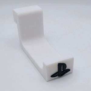 May include: A white, angled stand with a black PlayStation logo on the base. The stand is designed to hold a gaming controller. The design is simple and modern, with a curved shape and a small notch.