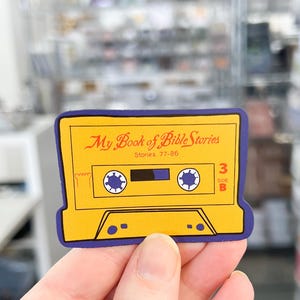 May include: Yellow and blue cassette tape sticker with the text "My Book of Bible Stories" and "Stories 77-85". The sticker has a blue border and a white cassette tape design.