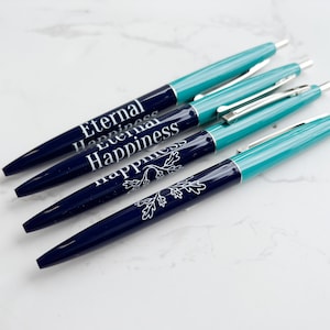 Puede incluir: Cuatro bolígrafos con barriles azul marino y turquesa están dispuestos en diagonal. Los bolígrafos tienen clips y puntas plateadas. El texto "Eternal Happiness" está impreso en un bolígrafo, y un diseño floral en otro.