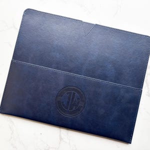 以下が含まれることがあります： V字型の切り込みが入ったフラップ付きのネイビーブルーの長方形ドキュメントフォルダー。フォルダーの前面には、JWの文字が入った円形のエンボス加工ロゴが表示されています。表面は滑らかでマットな質感で、縁にはステッチが施されています。