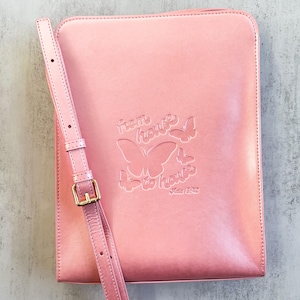 Könnte beinhalten: Eine rosa Leder-Umhängetasche mit einer goldenen Schnalle und einem Schmetterlingsdesign. Die Tasche hat den Text "from house to house" auf der Vorderseite geprägt.