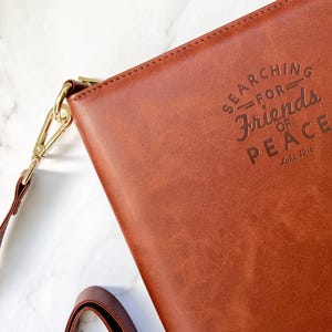 Puede incluir: Un bolso de cuero marrón con bandolera, cierre de hebilla dorado y un logotipo dorado en relieve que dice "SEARCHING FOR FRIENDS OF PEACE, est. 1976".