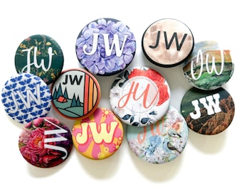 May include: A collection of 11 round buttons with various designs featuring the initials "JW" in different fonts and styles. The buttons have a variety of backgrounds, including floral patterns, landscapes, and abstract designs.