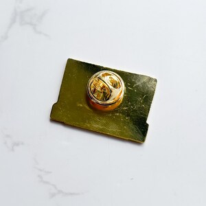 May include: A gold-colored rectangular pin with a round, gold-colored clasp on the back. The pin has a slightly distressed finish, giving it a vintage appearance. The pin is set against a white marble surface.