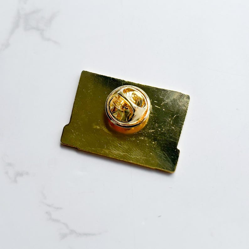 May include: A gold-colored rectangular pin with a round, gold-colored clasp on the back. The pin has a slightly distressed finish, giving it a vintage appearance. The pin is set against a white marble surface.
