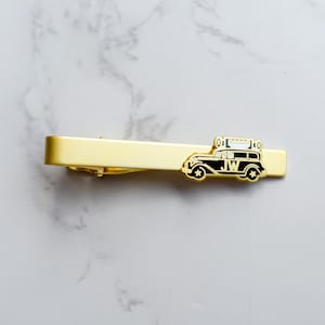 May include: A gold-colored tie clip with a black and white vintage car design. The car has the initials "JW" on the side. The tie clip is rectangular and has a shiny finish.