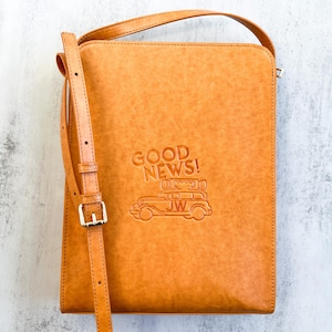 May include: A tan leather crossbody bag with a long adjustable strap. The bag has a flap closure and a embossed design of a car with the words "GOOD NEWS!" above it.