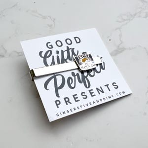 May include: A silver tie clip with a small, square, enamel charm that says "Trust in Jehovah". The charm has a yellow sun, blue mountains, and white clouds. The tie clip is on a white card with the text "Good Gifts Perfect Presents" and the website "GingerFiveAndDime.com".