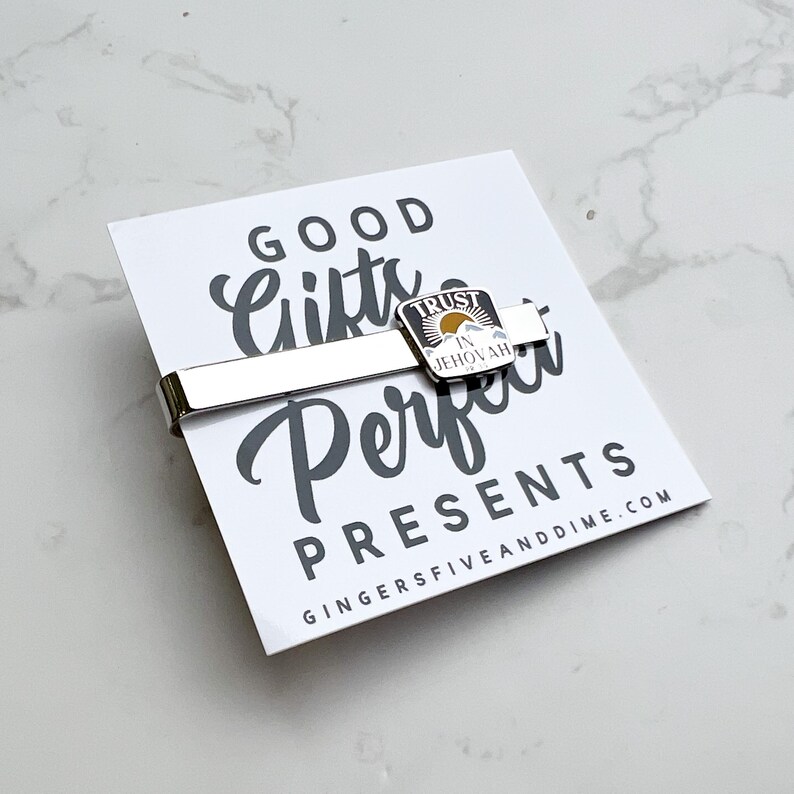 May include: A silver tie clip with a small, square, enamel charm that says "Trust in Jehovah". The charm has a yellow sun, blue mountains, and white clouds. The tie clip is on a white card with the text "Good Gifts Perfect Presents" and the website "GingerFiveAndDime.com".
