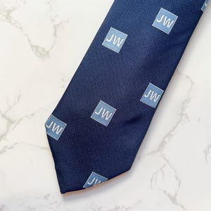 May include: Navy blue tie with a repeating pattern of light blue squares, each containing the white letters "JW". The tie is angled across a white and gray marble surface.