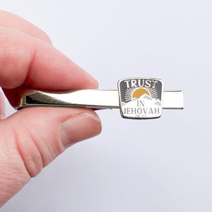 May include: Silver tie bar with a black and white enamel design. The design features a mountain range with a sun rising behind it. The text "TRUST IN JEHOVAH PS 33" is printed on the design.