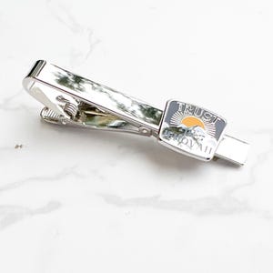 May include: Silver tie bar with a rectangular enamel charm that says "TRUST THE JOURNEY". The charm has a yellow sun and a mountain range design.