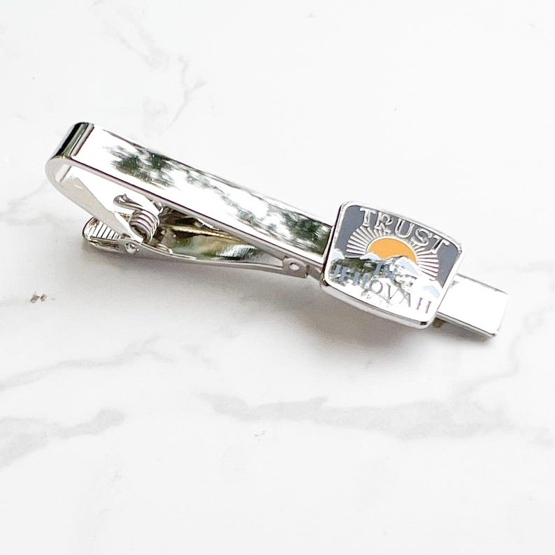 May include: Silver tie bar with a rectangular enamel charm that says "TRUST THE JOURNEY". The charm has a yellow sun and a mountain range design.