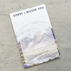 May include: A rectangular card with the text "SORRY I MISSED YOU" at the top. The card features a watercolor landscape of mountains and a valley. The design incorporates soft blues, purples, and greens.