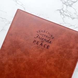 Puede incluir: Un diario de cuero marrón con el texto en relieve "SEARCHING FOR FRIENDS OF PEACE" y el versículo bíblico Lucas 10:6. El diario tiene un borde cosido y está sobre un fondo de mármol.