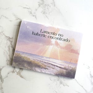 Könnte beinhalten: Eine rechteckige Karte mit einem Aquarellgemälde eines Sonnenuntergangs über dem Meer. Der Himmel wechselt von Lila zu Rosa, wobei die Sonnenstrahlen durch die Wolken brechen. Der Text "Lamento no haberle encontrado" ist auf die Karte gedruckt.