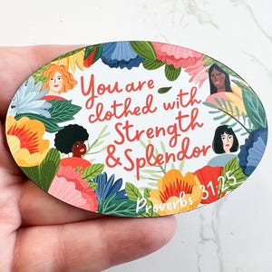 May include: Oval-shaped wooden sign with a floral border and three women's faces. The sign reads "You are clothed with Strength & Splendor Proverbs 31:25".