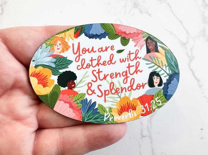 May include: Oval-shaped wooden sign with a floral border and three women's faces. The sign reads "You are clothed with Strength & Splendor Proverbs 31:25".