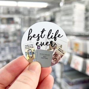 May include: A white round sticker with black text that says "best life ever." The sticker has a collage of images of books and a phone with a blue screen. The sticker is being held by a hand.