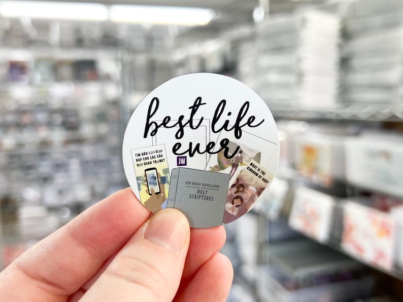 May include: A white round sticker with black text that says "best life ever." The sticker has a collage of images of books and a phone with a blue screen. The sticker is being held by a hand.