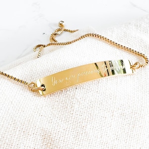 May include: Gold-colored bracelet with a rectangular plate engraved with the words "You are precious to Jesus." The bracelet has a delicate chain and a sliding clasp for adjustable sizing. The bracelet is resting on a white surface.