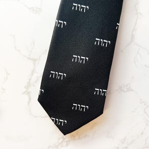 May include: A black necktie with a white Hebrew word pattern. The word is repeated throughout the tie.
