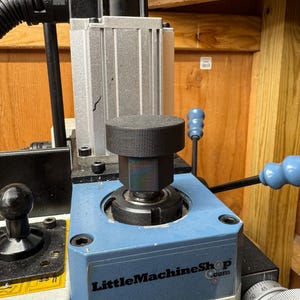 May include: A close-up of a blue and silver mini mill machine. The machine has a black knob on top and blue handles. The text "LittleMachineShop.com" is visible on the machine.