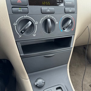 May include: A car's dashboard with a digital clock displaying 12:55, a black and gray control panel with buttons and knobs, and a black gear shift lever with a 'P' for park.