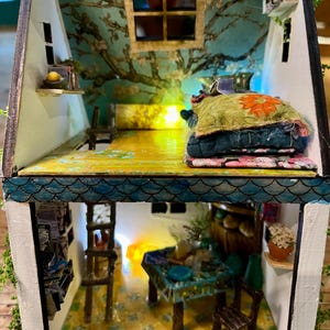 May include: A two-story dollhouse with a blue tree-patterned wall, a wooden ladder, and miniature furniture. The upper level features a bed with stacked floral pillows, while the lower level contains a table, chairs, and a small potted plant.