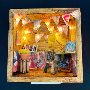 May include: A miniature wooden room with a desk, chair, and bookshelves. The room is decorated with string lights, paper flags, and a small potted plant. The walls are covered in patterned paper.