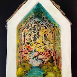 May include: A miniature cardboard house with a painted scene of a forest and a stream inside. The house is white with a brown cardboard roof. The scene inside is painted in shades of green, blue, and brown. There is a small stream with a waterfall in the center of the scene. The forest is painted in the background.
