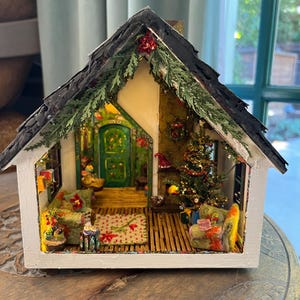 May include: A miniature Christmas-themed house with a white exterior, black shingled roof, and a detailed interior. The interior features a Christmas tree, furniture, and decorative elements. The house is decorated with greenery and festive accents.
