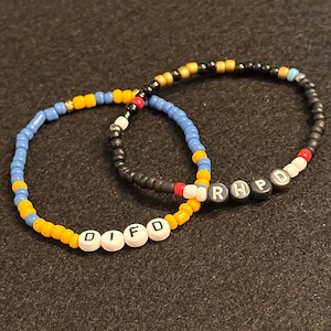 May include: Two beaded bracelets with the names "DIFD" and "RHPD" spelled out in white beads on black beads. The bracelets are made with yellow, blue, black, white, and red beads.