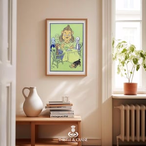 May include: A framed print featuring a colorful illustration of characters from The Wizard of Oz, including the Cowardly Lion, Dorothy, and the Tin Man, gathered around a table. The print is displayed on a light-colored wall above a wooden table with books and a white vase.