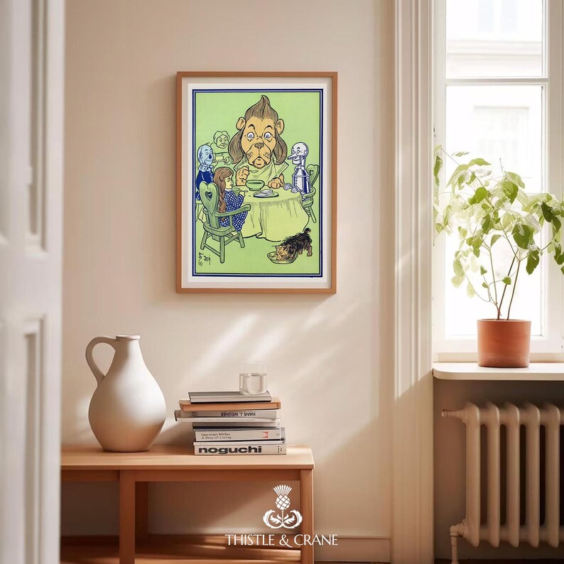 May include: A framed print featuring a colorful illustration of characters from The Wizard of Oz, including the Cowardly Lion, Dorothy, and the Tin Man, gathered around a table. The print is displayed on a light-colored wall above a wooden table with books and a white vase.