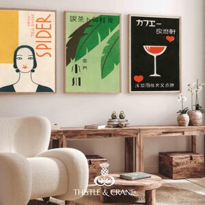 May include: Three framed vintage art prints. The first features a woman's portrait with the word "SPIDER". The second has green leaves and Japanese text. The third has a cocktail glass and Japanese text. The prints are displayed above a wooden console.