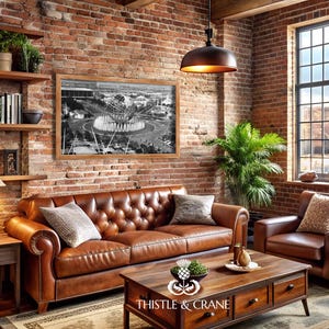 May include: A brown leather tufted sofa and matching armchair in a room with exposed brick walls. A wooden coffee table features a Thistle & Crane logo. A framed black and white photograph of the Unisphere hangs above the sofa near a window.