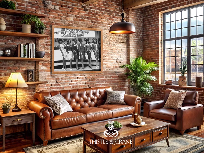 May include: A framed black and white photograph featuring six women standing in a row with the text DU PONT DAY PRETTIEST LEGS AT THE FAIR CANTRECE NYLON displayed above them. The print is mounted on a brick wall above a brown leather sofa.