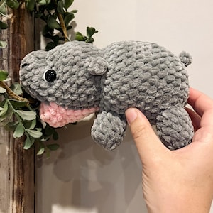 May include: A gray crocheted plush hippopotamus toy with pink detailing around the mouth. The toy has black eyes and is being held by a hand.
