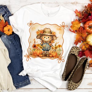 May include: White t-shirt featuring a watercolor illustration of a scarecrow with a hat, surrounded by pumpkins and sunflowers. The design is framed with a decorative border. The shirt is paired with blue jeans and leopard print flats, evoking a fall theme.