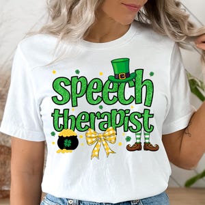Puede incluir: Camiseta blanca con temática del Día de San Patricio. La camiseta presenta las palabras "Speech therapist" en letras verdes brillantes, un sombrero de duende, una olla de oro, un lazo amarillo y piernas y zapatos de duende.