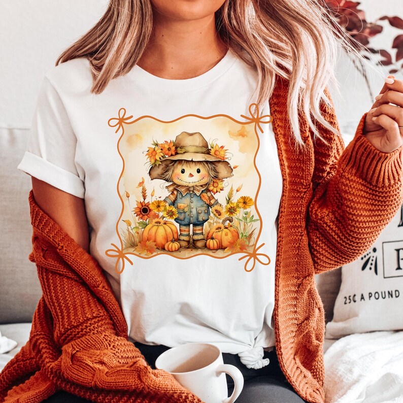 May include: White t-shirt featuring a watercolor illustration of a scarecrow with sunflowers, pumpkins, and autumn foliage. The scarecrow wears a hat and overalls. The design is framed with an orange border, perfect for fall.