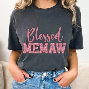 K&ouml;nnte beinhalten: Ein dunkelgraues T-Shirt mit dem Text "Blessed Memaw" in pinkfarbenem Glitzer.