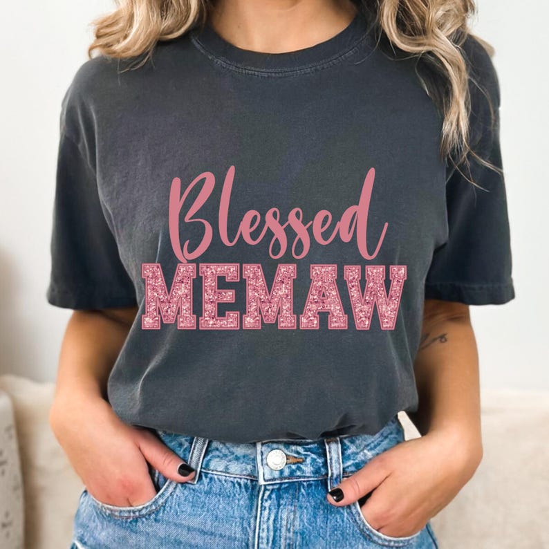 K&ouml;nnte beinhalten: Ein dunkelgraues T-Shirt mit dem Text "Blessed Memaw" in pinkfarbenem Glitzer.