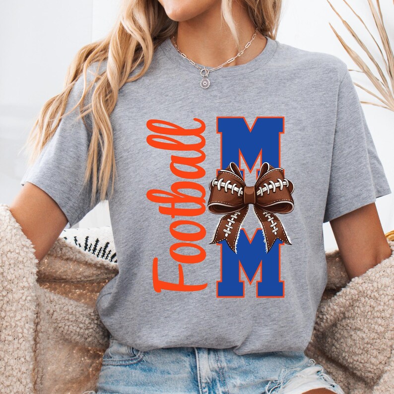 May include: A gray t-shirt with the word "Football" in orange script on the left side. The shirt features the word "MOM" in blue block letters, with a football-shaped bow in the center. The bow is brown with white laces.
