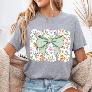 Op de afbeelding: Een grijs t-shirt met een grote, lichtgroene strik. De strik staat boven een rechthoekig ontwerp met een bloemenpatroon van roze, oranje en groene bloemen en bladeren. Het t-shirt heeft een casual, comfortabele stijl.