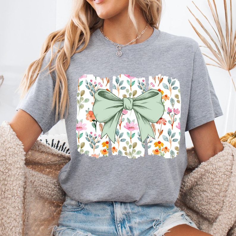 Op de afbeelding: Een grijs t-shirt met een grote, lichtgroene strik. De strik staat boven een rechthoekig ontwerp met een bloemenpatroon van roze, oranje en groene bloemen en bladeren. Het t-shirt heeft een casual, comfortabele stijl.