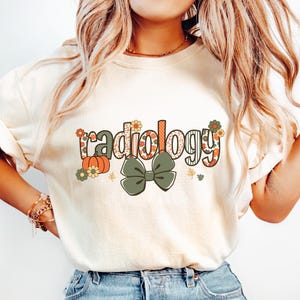 May include: Cream-colored t-shirt featuring the word "radiology" in a floral and patterned design. A green bow and fall-themed accents, including pumpkins and leaves, are also present. The shirt is a crew neck style.