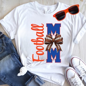 May include: White t-shirt with the text "Football Mom" in orange script. The word "MOM" is in blue block letters with a brown football-shaped bow in the center. Orange sunglasses rest on the shirt. Blue jeans and white sneakers are also visible.
